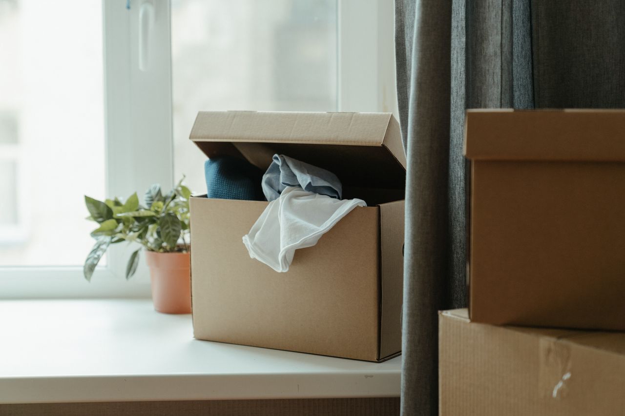An open cardboard box filled with clothes beside a window, representing a move or relocation.